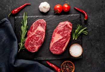 Two raw marbled beef ribeye steaks with spices on a stone background