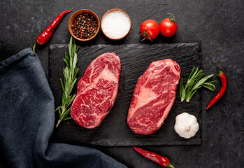 Two raw marbled beef ribeye steaks with spices on a stone background
