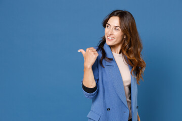Smiling cheerful funny pretty beautiful young brunette woman 20s wearing basic jacket standing pointing thumb aside on mock up copy space isolated on bright blue colour background, studio portrait.