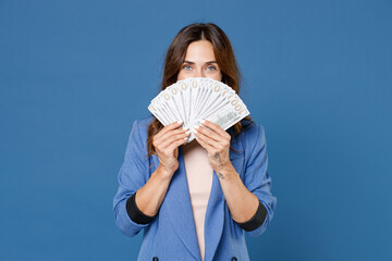 Cheerful young brunette woman 20s wearing basic jacket standing covering face with fan of cash money in dollar banknotes looking camera isolated on bright blue colour wall background studio portrait.