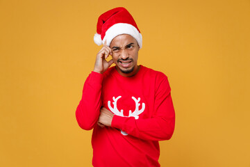 Puzzled young Santa african american man in red sweater Christmas hat put hand on head looking aside isolated on yellow background studio portrait. Happy New Year celebration merry holiday concept.