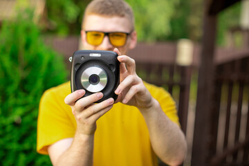 Young happy male photographer in yellow T-shirt and trendy sunglasses having fun outdoors, takes photo with instant camera on green nature background, resting time. Summer, leisure, hobby concept