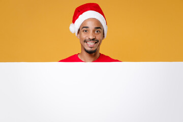 Smiling young Santa african american man in Christmas hat hold big white empty blank billboard mock up copy space isolated on yellow background studio. Happy New Year celebration holiday concept.
