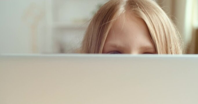 Cute schoolkid hides behind laptop with smiling face.Close up female eyes looking from screen of computer. Portrait of funny small teen girl play at table doing homework online in net, happy chilhood