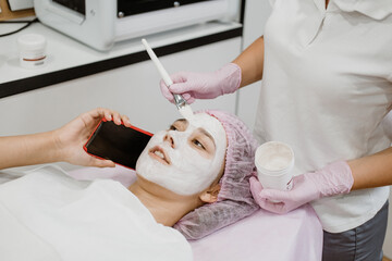Obraz na płótnie Canvas Professional skin care in beauty salon. Young woman with clay face mask takes selfie on phone. Facial care by beautician at spa salon. Acne Treatment, face peeling mask, spa beauty treatment