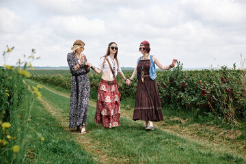 Three young hippie women, wearing boho style clothes, walking holding hands, dancing on green currant field in summer. Eco tourism concept. Friends traveling in rural countryside.