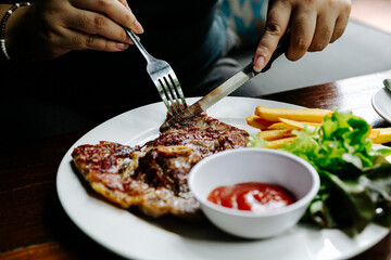 eating steak on white plate with fork and knife on his hands