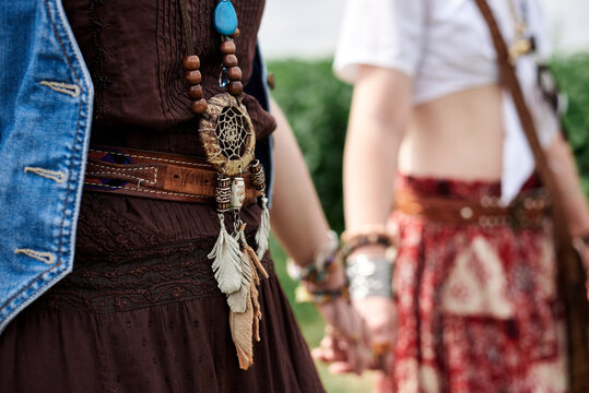 Close-up picture of hands with colorful bracelets and various rings, holding each other. Hippie women, wearing boho style clothes, standing on green field. Female friendship concept.