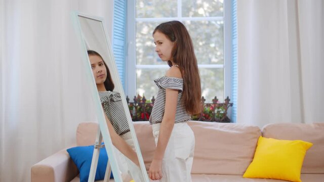Side view of pretty preteen girl in casual wear looking in full size mirror and smiling