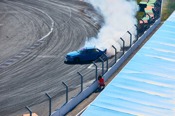 A blue race car drifts powerfully, producing a large cloud of white smoke on a gray asphalt track during a daytime race event for automotive or sports promotions. © LemPro Filming Life
