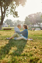 Happy lesbian couple of women looking at each other while sitting on the grass in summer park, spending time together, hugging each other