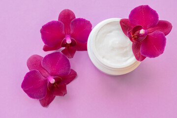 White container with cream for face and body with magenta orchid flowers on pink background. Concept of delicate facial cosmetics with orchid extract