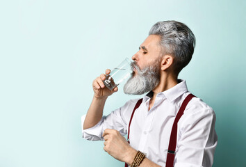 Gray-haired elderly male in white shirt, brown suspenders. He drinking pure water from glass, posing sideways on blue background