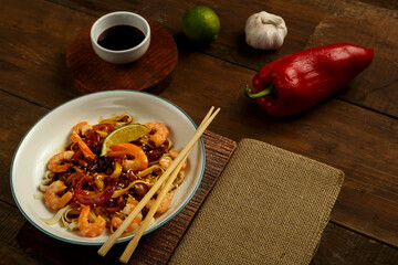 Close-up of udon with vegetables, mushrooms and shrimps in a light plate near sticks.