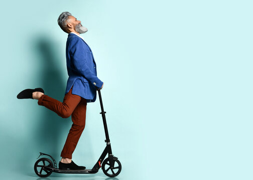 Gray-haired Male In White Shirt, Jacket, Brown Pants And Loafers. Riding Black Scooter And Posing Sideways On Blue Background