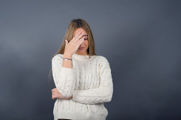 Beautiful young blonde woman wearing white sweater standing against gray background, 