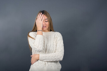 Beautiful young blonde woman wearing white sweater standing against gray background, 