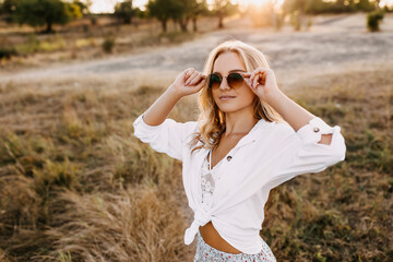Young woman with blond hair posing outdoors, wearing round sunglasses.