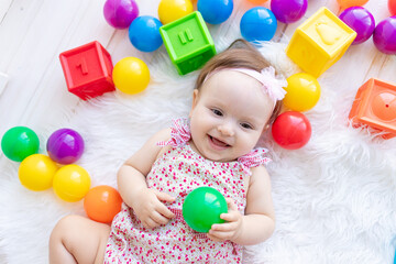 Fototapeta na wymiar a beautiful little baby girl is lying in red clothes on a white Mat among toys balls and cubes