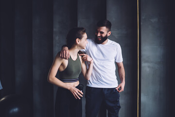 Positive caucasian female and male best friends having conversation after workout in gym enjoying healthy lifestyle, smiling hipsters in tracksuit having fun and joking on break during training.