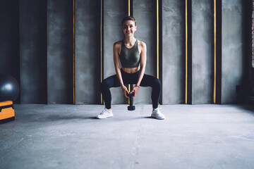 Positive caucasian female in active wear squatting with weight training lower body muscles during workout, smiling woman looking at camera squatting with dumbbells for slimming on healthy lifestyle