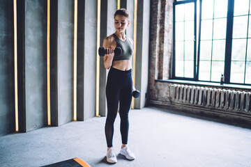 Determined caucasian female in active wear standing in gym working on arms muscles holding dumbbells, serious pretty strong professional sports women lifting weight training upper body biceps