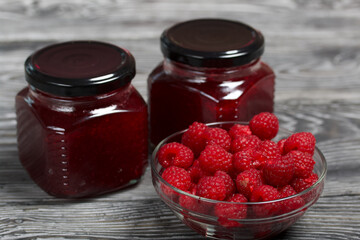 Raspberry jam in glass jars. Nearby are raspberries in a container. On wooden boards with a beautiful texture.