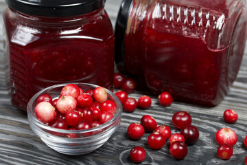 Cranberry jam in glass jars. Nearby berries are in a container and scattered over the surface. Close-up. On wooden boards with a beautiful texture.