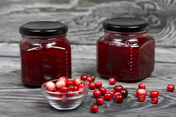 Cranberry jam in glass jars. Nearby berries are in a container and scattered over the surface. On wooden boards with a beautiful texture.