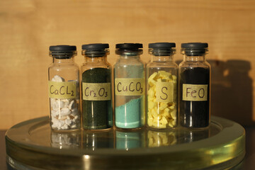 Various inorganic crystalline substances in glass jars with the formulas: white calcium chloride, green chromium oxide, blue copper carbonate, yellow sulfur, black iron oxide.