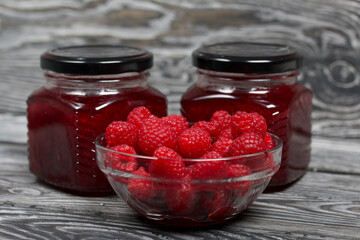 Raspberry jam in glass jars. Nearby are raspberries in a container. On wooden boards with a beautiful texture.