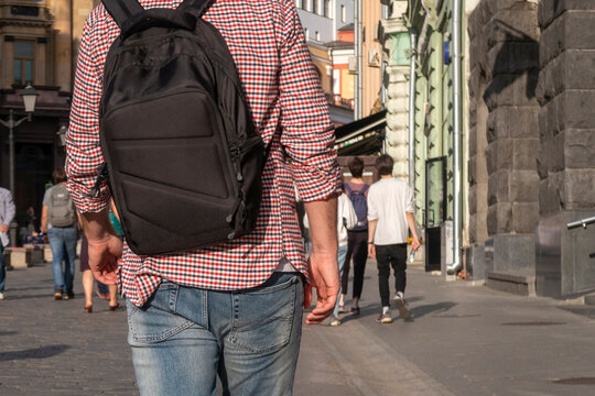 Man's back with a black backpack, wearing blue jeans and a plaid shirt. City street with walking people on sunny afternoon.