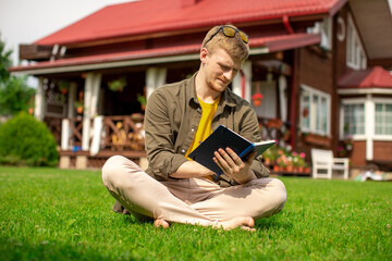 barefoot young man wearing yellow glasses sitting on green grass making notes in his notebook. Country house on blurred background. beginner writer writes essay on sunny summer day