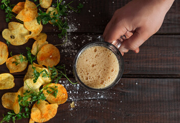 Men's hand holding a glass with dark beer on dark rustic wooden table with homemade potato chips with parsley. Tasty fast food. Backdrop of the Oktoberfest. Top view. Flat lay