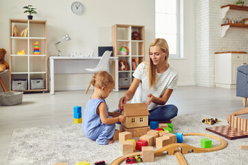 Young mom playing with cute little son and teaching him to build toy house in cozy modern apartment - obrazy, fototapety, plakaty