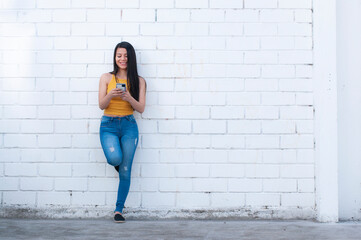 full body photo of beautiful latina woman texting on cell phone leaning against a white wall - obrazy, fototapety, plakaty