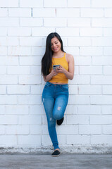 beautiful latina woman writing on her cell phone leaning against a white wall