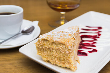 A slice of Napoleon cake, served with a cup of coffee and a glass of brandy. Cafe decoration and dessert concept. Selective focus