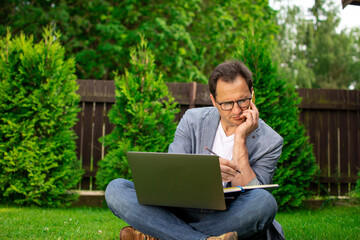 Multitasking. Middle aged businessman freelancer sits outdoors on lawn holds laptop, talks on mobile phone and makes notes in notebook. Busy vacation, coffee break, overworked, small business support