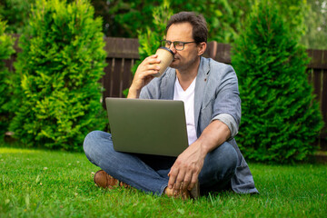 Attractive middle aged businessman resting outdoors sits on grass with coffee to-go and laptop, office worker have coffee break, male freelancer works in park, pause, relax, leisure concept