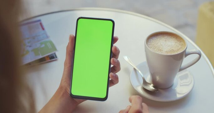 Crop view of female person holding smartphone and swipping greencreen while sitting at table with coffee and map on it. Concept of mock up screen and chroma key.