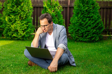 Sad and upset unshaven mature European 40s businessman sits on grass with laptop. dramatic...