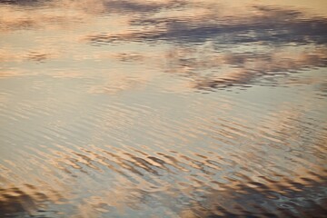 Shiny surface of calm water with golden highlights and reflections. Beautiful glowing ripples on water surface close-up. Water surface with abstract pattern and white clouds and blue sky reflections. 