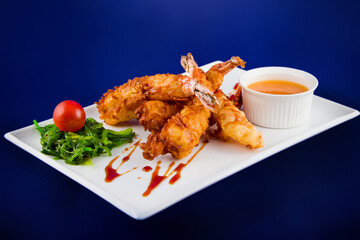 Deep-fried tiger prawns in batter served with sweet and sour sauce, nori seaweed and cherry tomatoes on a rectangular plate.