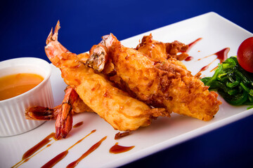 Deep-fried tiger prawns in batter served with sweet and sour sauce, nori seaweed and cherry tomatoes on a rectangular plate.