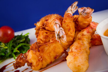 Deep-fried tiger prawns in batter served with sweet and sour sauce, nori seaweed and cherry tomatoes on a rectangular plate.