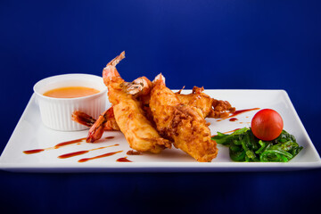 Deep-fried tiger prawns in batter served with sweet and sour sauce, nori seaweed and cherry tomatoes on a rectangular plate.