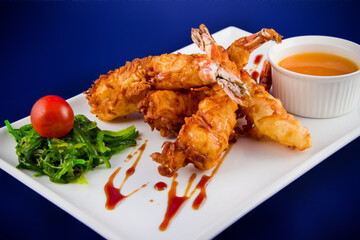 Deep-fried tiger prawns in batter served with sweet and sour sauce, nori seaweed and cherry tomatoes on a rectangular plate.