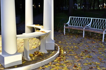 Evening. Illuminated gazebo with white columns. Not far from it is an empty bench. The paths are...