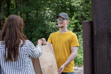 Handsome friendly deliver man in yellow uniform handing paper bags with food, gives order to female customer in front of country house. Express grocery delivery service, online shopping